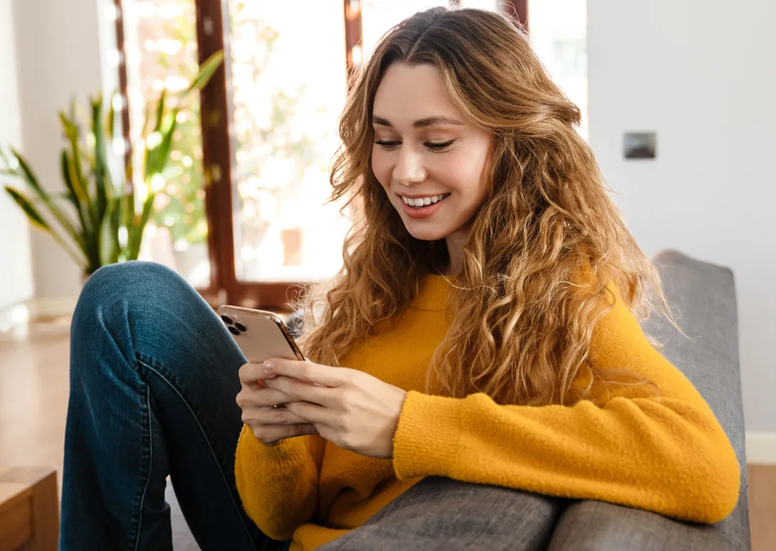 Smiling woman with wavy hair wearing a yellow sweater, sitting on a couch and looking at her smartphone in a bright living room.